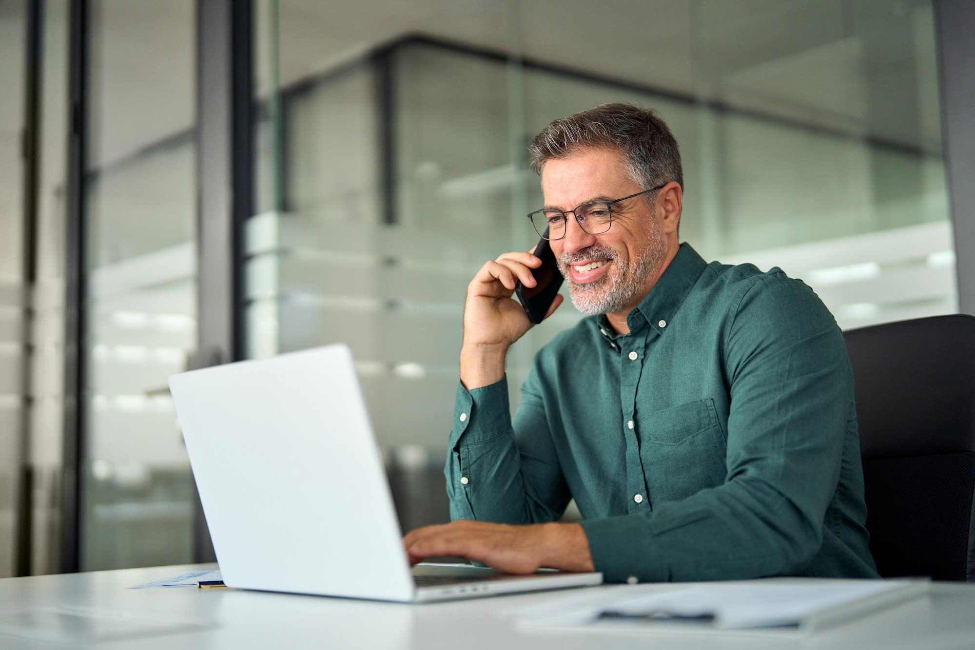Busy smiling older business man making phone call using laptop in office.
